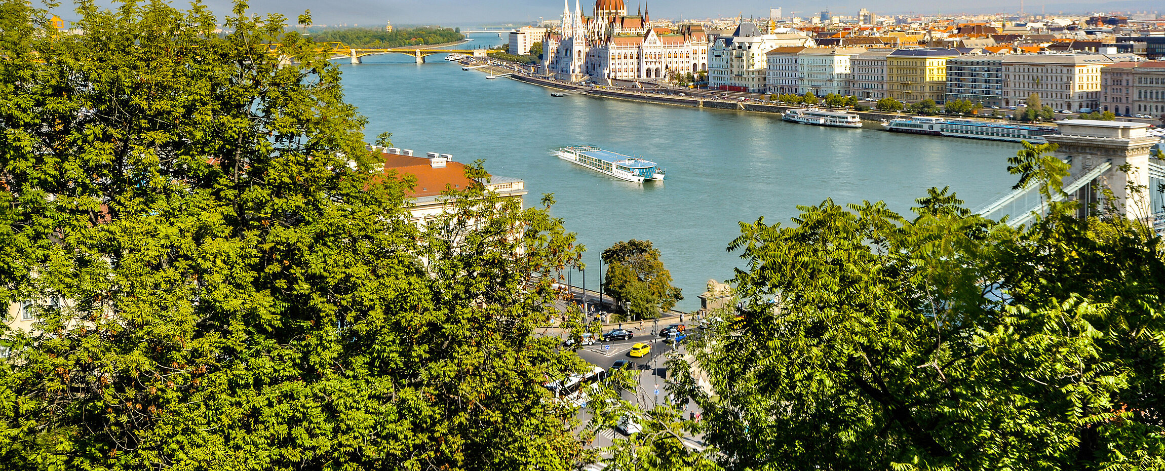 Luftaufnahme der Donau in Budapest mit Blick auf das ungarische Parlamentsgebäude und die Kettenbrücke, umrahmt von grünen Baumwipfeln.