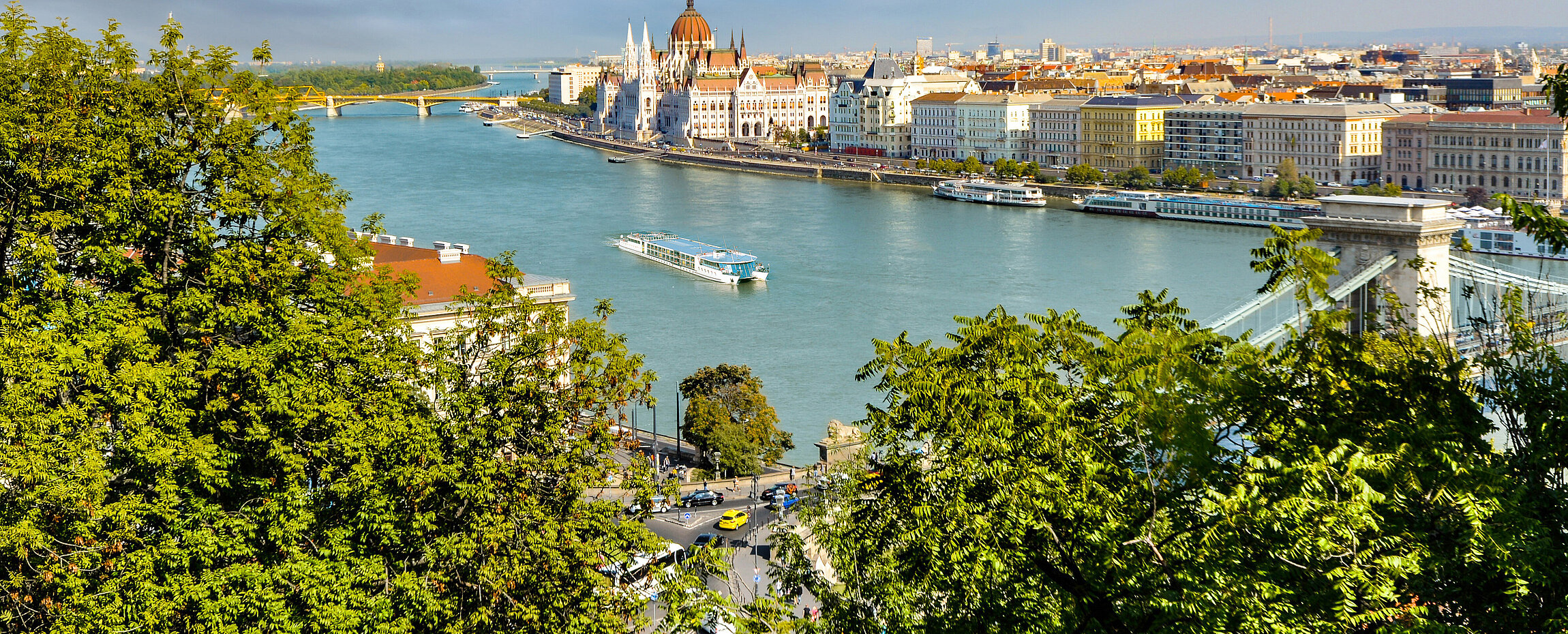 Luftaufnahme der Donau in Budapest mit Blick auf das ungarische Parlamentsgebäude und die Kettenbrücke, umrahmt von grünen Baumwipfeln.