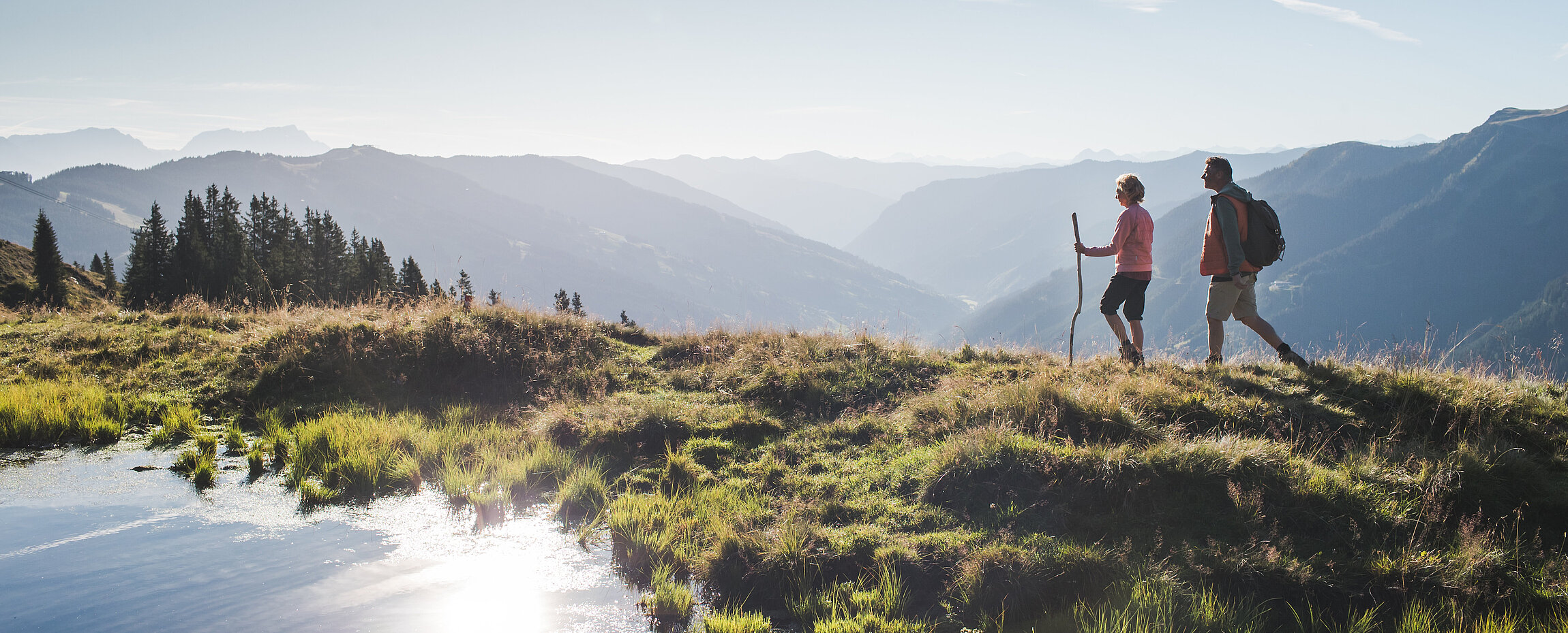 Wanderpaar am Berg mit kleinem Bergteich, dahinter Tallandschaft.