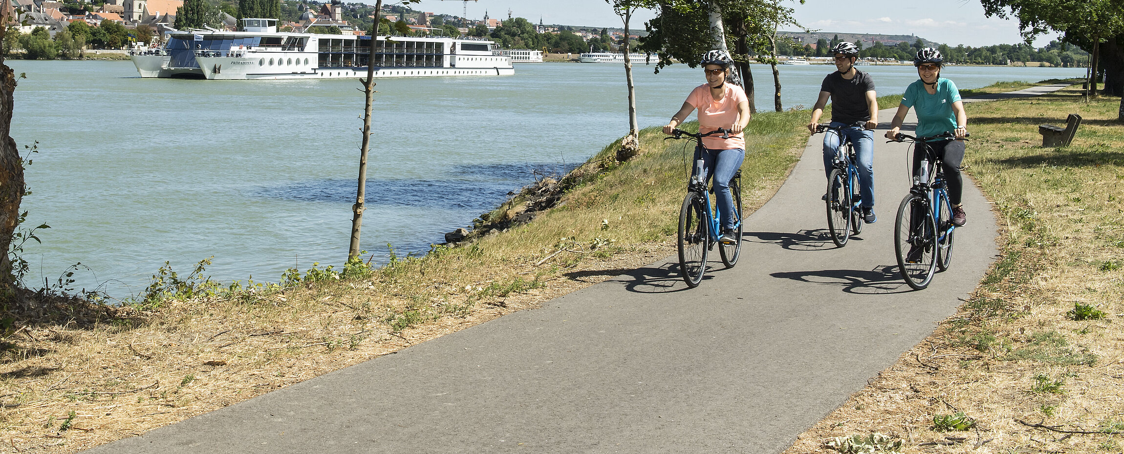 drei Radler fahren entland der Dona, ein Schiff fährt im Hintergrund